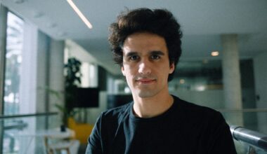 A man wearing a large black t-shirt stands in a sunlit office.