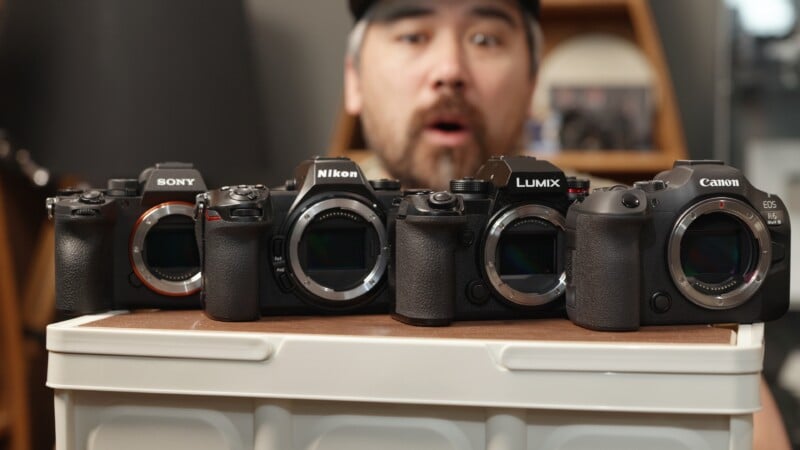 A man with a surprised expression stands behind four mirrorless cameras—Sony, Nikon, Lumix, and Canon—lined up on a white surface, with all camera lens mounts visible.
