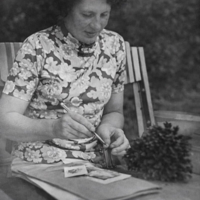 A woman in a floral shirt sits outdoors on a striped chair, painting with a brush. She appears focused on her work, with art supplies and a small plant or shrub in front of her. The image is black and white.
