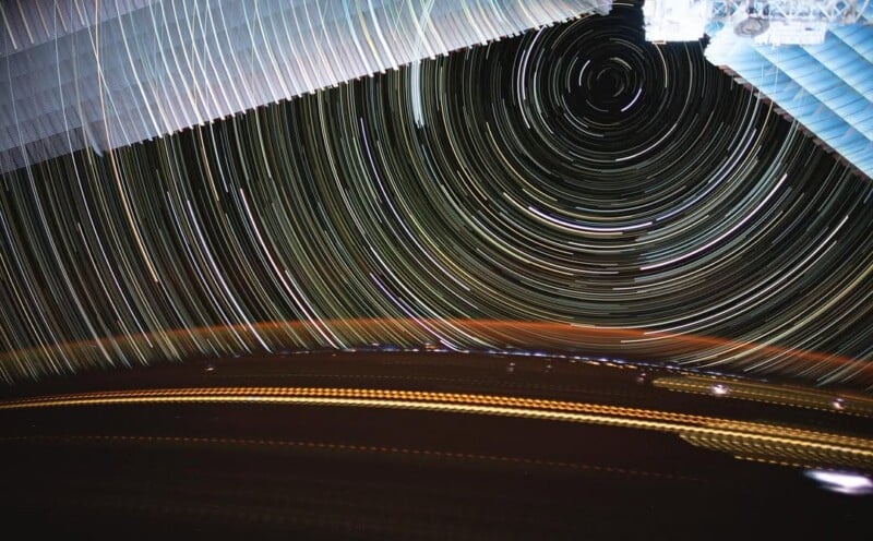 Long-exposure photo showing colorful circular star trails in the night sky, with part of a space station’s solar panels visible at the top and blurred lights across the bottom.