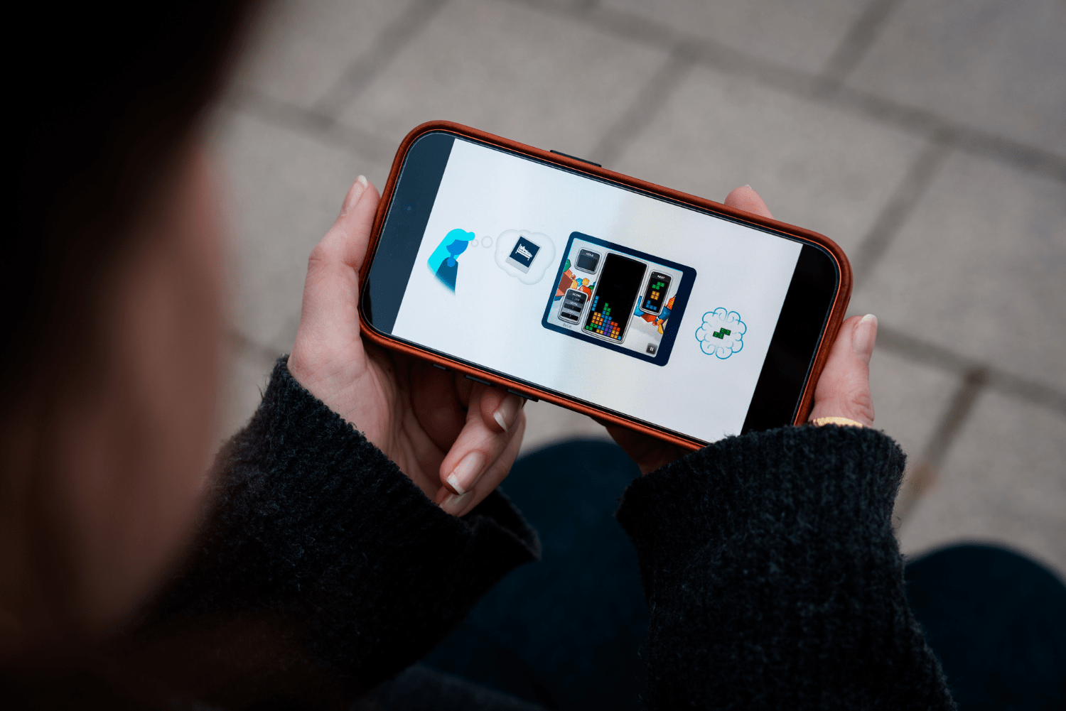 A phone held in a woman's hands shows an illustration of a Tetris game next to a cartoon person with a thought bubble