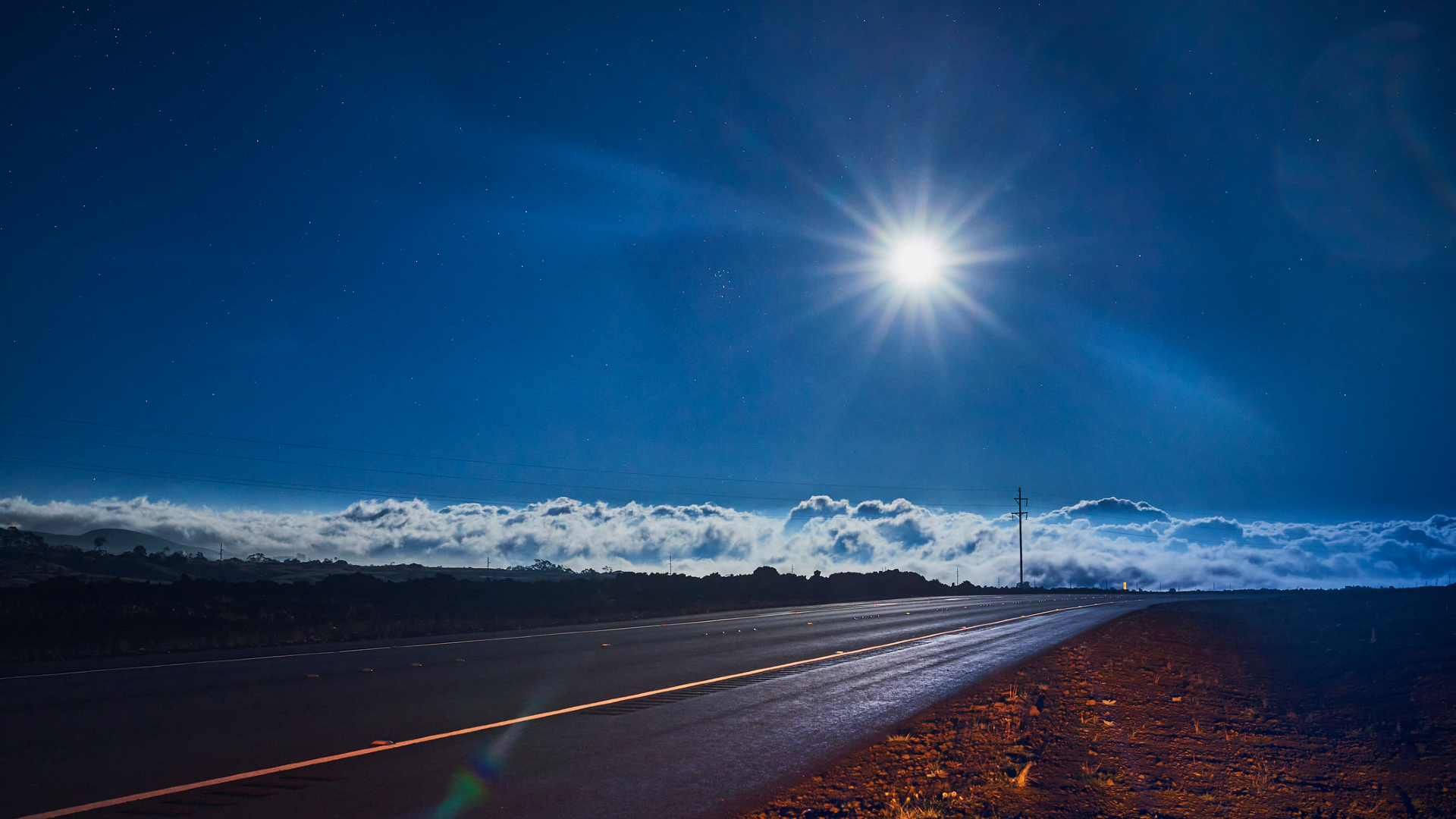 A bright sun in a blue sky looks down on a surface of clouds in the background while a road comes toward the bottom left of the image in the foreground