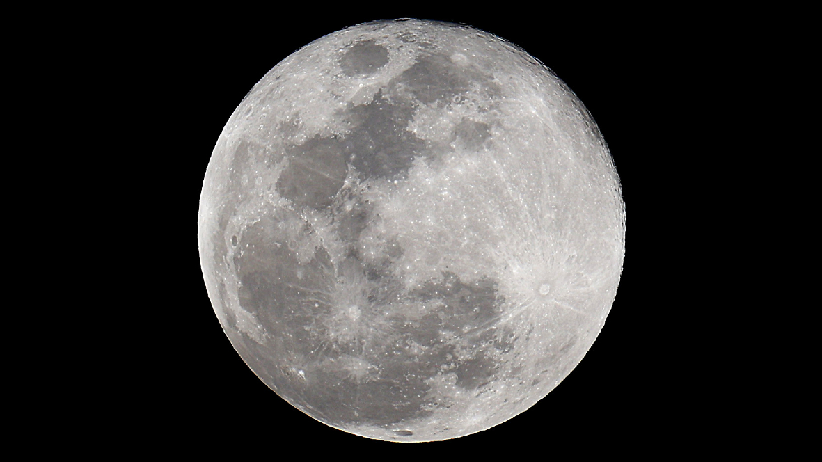 A silvery full moon is pictured against a black night sky. The dark basalt plains of lunar maria are visible covering swathes of its surface. Bright streaks of material are also visible emanating from prominent craters.