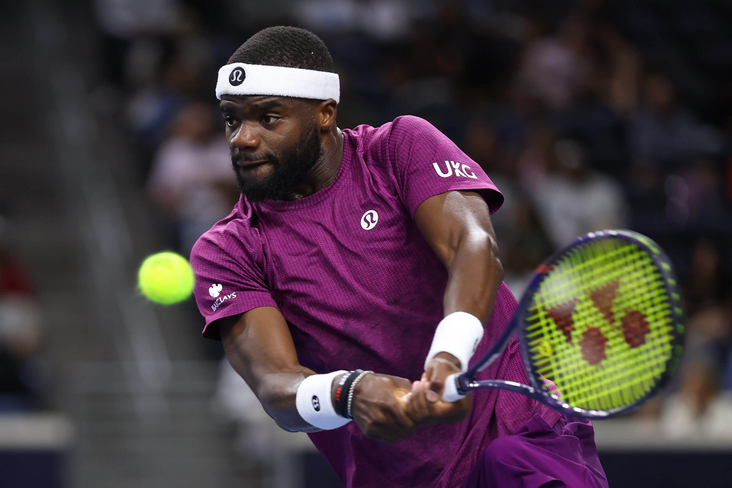 Frances Tiafoe hits a two-handed backhand wearing a purple t-shirt with a UKG logo on his left sleeve and a Barclays logo on his right sleeve.