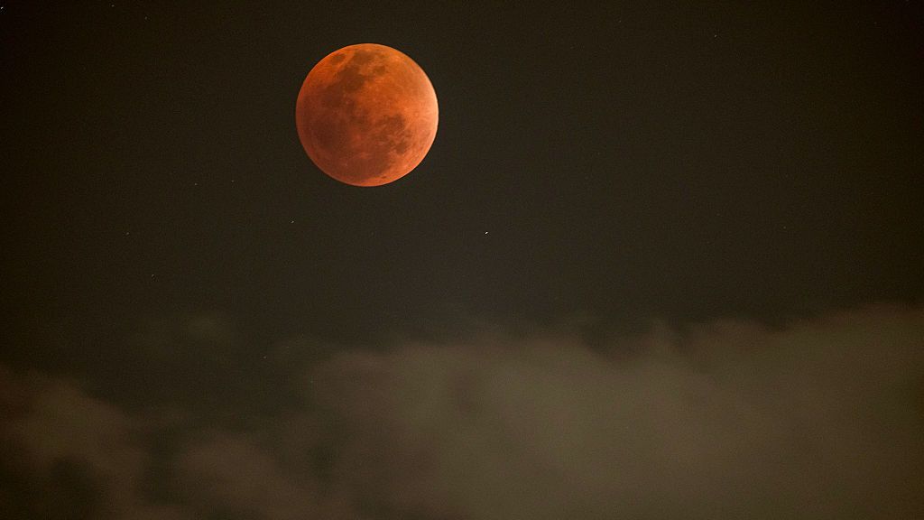 A red full moon is pictured above grey clouds in a dark night sky.