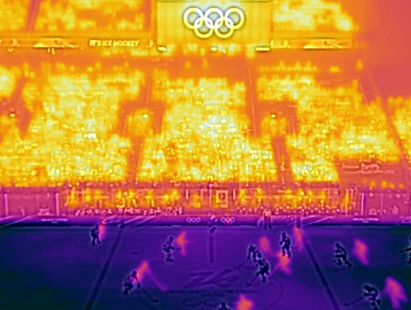 Thermal image of an Olympic ice hockey game, showing bright yellow and orange heat signatures from the audience and cooler purple and blue tones on the ice and players. The Olympic rings are visible above the crowd.