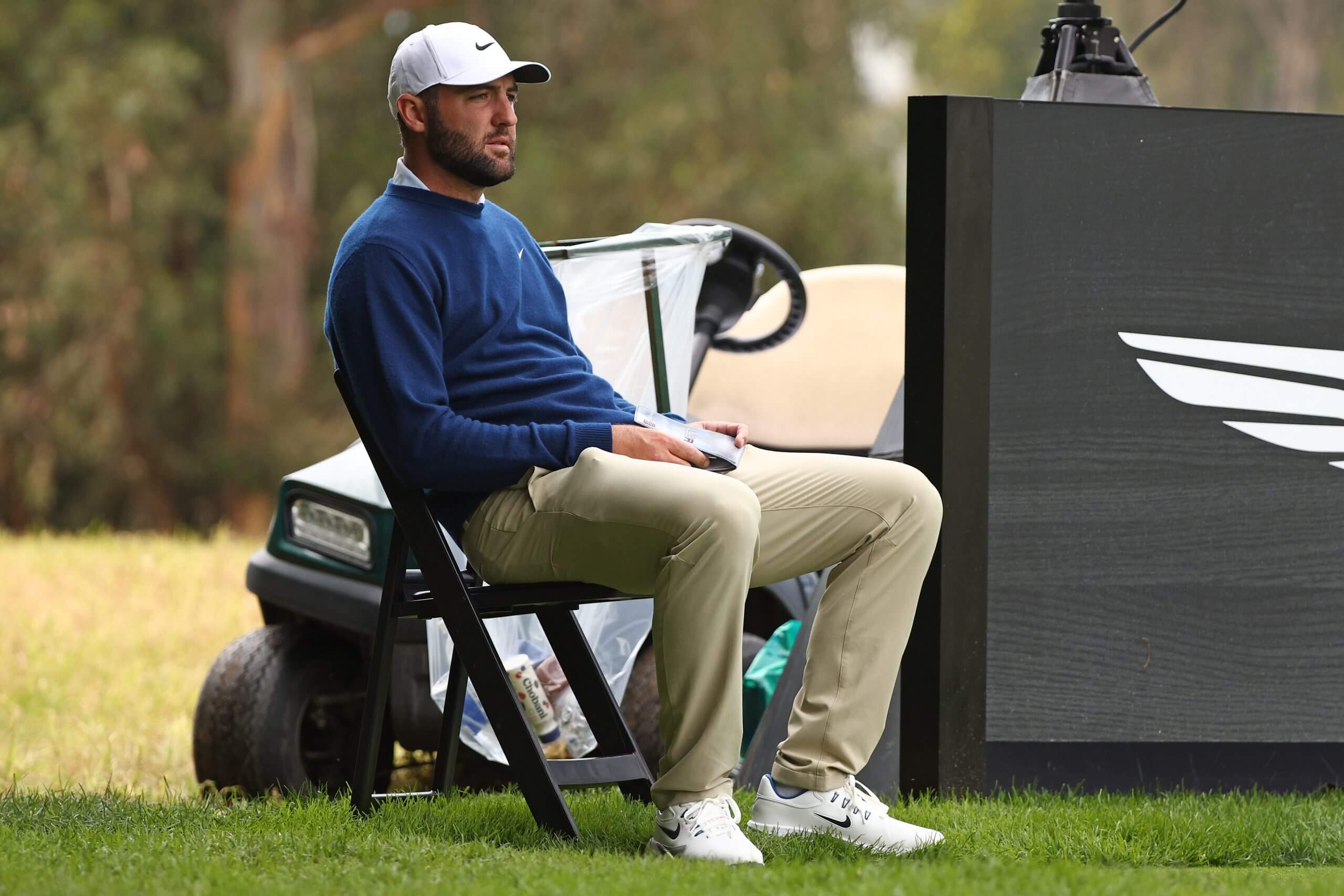 Scottie Scheffler — in khaki pants, a blue sweater and white hat — sits in a chair on the eighth hole.