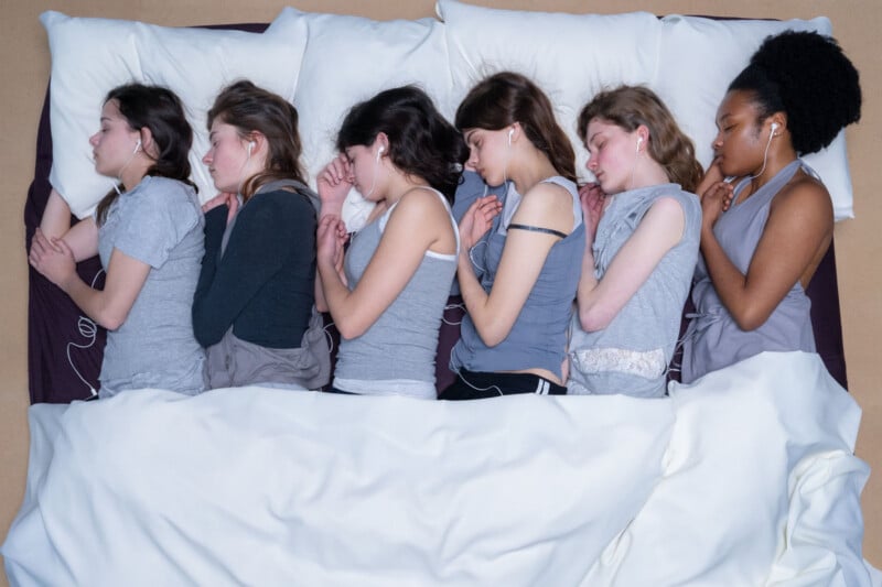 Six young women lie side by side in bed, facing the same direction, with eyes closed and earbuds in. They are under a white blanket with white pillows, wearing similar gray sleepwear.