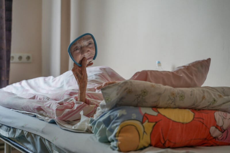 An elderly person lies in bed, covered by blankets, holding a small mirror to look at their reflection. The room is softly lit and there are colorful pillows beside them.