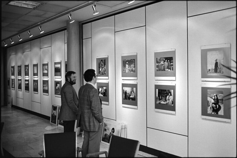 Two men stand in a gallery, closely viewing black-and-white photographs displayed on a white-paneled wall. More framed photos line the walls, creating an exhibition-like setting with overhead lighting.