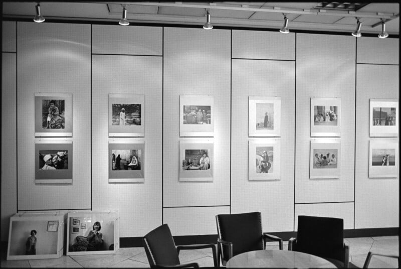 Black and white photo of a gallery wall displaying framed photographs in three rows, with three empty chairs and a round table in the foreground. Several unmounted photos rest on the floor against the wall.