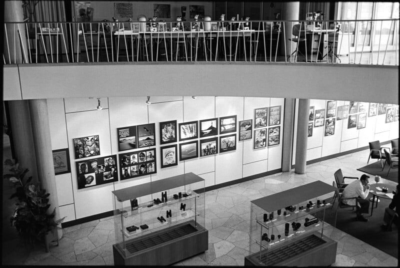 Black and white photo of an art gallery with framed artworks on the wall, display cases with objects, two people seated at a table, and an upper level with more artworks and desks visible above.