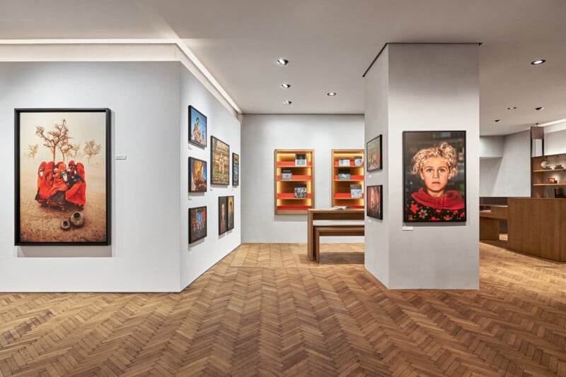 A modern art gallery with framed photographs on white walls, a wooden herringbone floor, illuminated shelves with books, and a wooden table in the center. Bright lighting highlights the artwork and cozy interior.