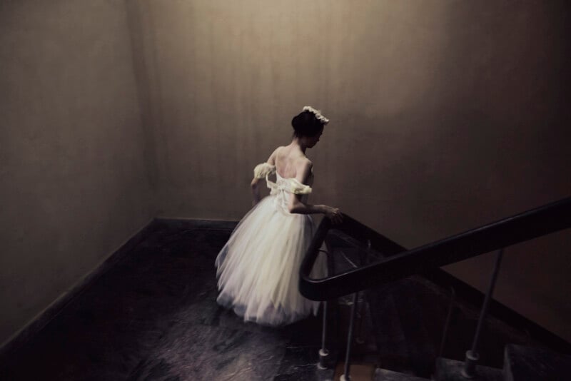A woman in a white, off-shoulder gown and floral headpiece descends a dark, winding staircase, her back turned to the camera, in a dimly lit, vintage-looking setting.