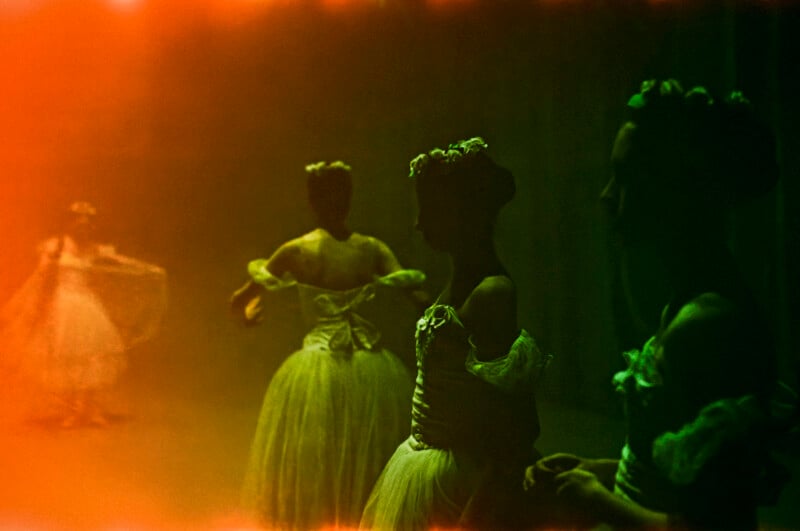 Three ballerinas in tulle dresses and floral headpieces stand in low light with a warm orange glow, creating a soft, dreamy, vintage effect. One dancer is in the background, while the others are in the foreground.