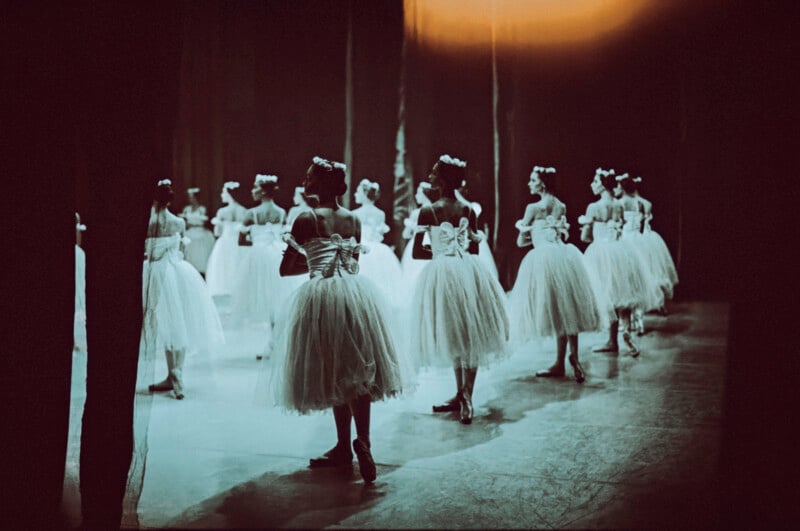 A group of ballerinas in white tutus with large bows stand on stage, viewed from behind and slightly to the side, in dim, dramatic lighting before a performance.