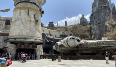 The Millennium Falcon is displayed in a theme park, surrounded by visitors. Rocky formations and sci-fi architecture provide a backdrop under a partially cloudy sky.