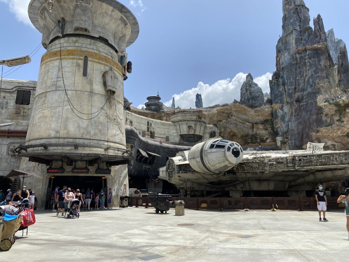 The Millennium Falcon is displayed in a theme park, surrounded by visitors. Rocky formations and sci-fi architecture provide a backdrop under a partially cloudy sky.