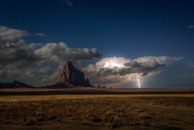 A dramatic landscape showing Shiprock, a tall rock formation under a cloudy night sky, as a bolt of lightning illuminates clouds in the distance over an arid, grassy plain.