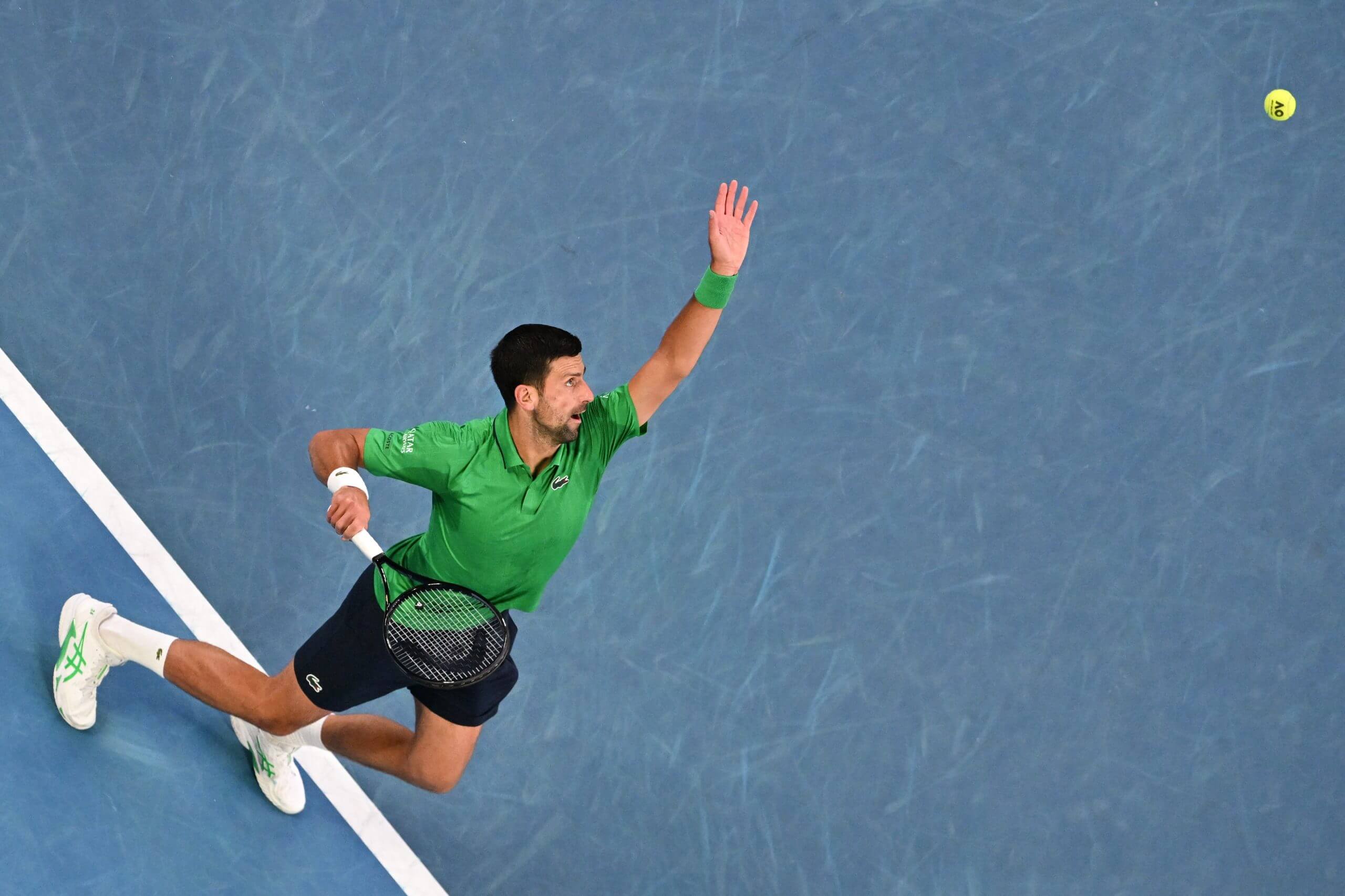 Novak Djokovic goes into the trophy position as he prepares to hit a serve on a blue tennis court.