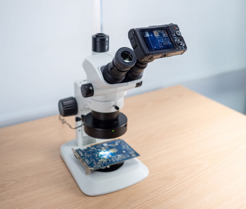 A white microscope with a digital camera attached is positioned on a light wooden table, focused on examining a blue circuit board placed on its base.