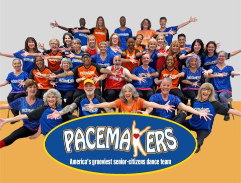 Image of a group of adults wearing blue and orange tshirts with arms open wide smiling at the camera behind a logo that reads Pacemakers