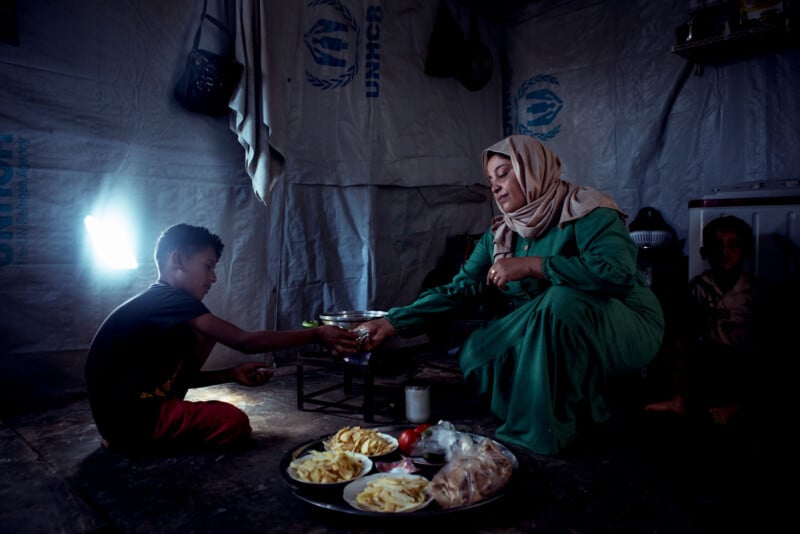 A woman in a green dress sits inside a UNHCR tent, handing food to a young boy. A tray with bread, fries, and vegetables is on the floor. The setting is dimly lit, highlighting a sense of intimacy and care.