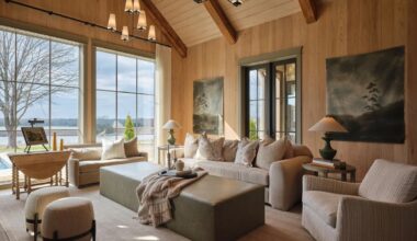 A cozy living room with textured, wood-paneled walls, large windows, warm interiors in neutral tones, two landscape paintings by Claire Thompson, and a view of trees and water outside.