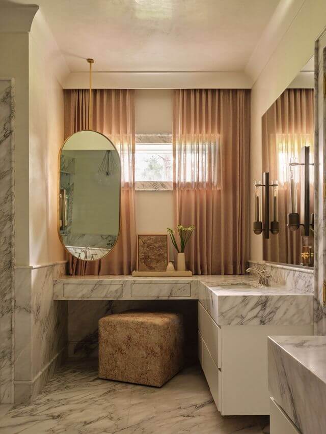 Claire Thompson's Interior Designs Are Soft, Warm, and Endlessly Livable - 12 A marble bathroom vanity with an oval mirror, beige stool, and pink curtains over two windows, featuring brass and gold accents.