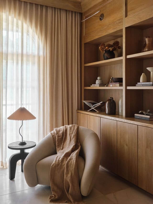 Claire Thompson's Interior Designs Are Soft, Warm, and Endlessly Livable - 13 A cozy reading nook with a round white chair draped with a beige throw, a small lamp on a side table, and built-in wooden shelves holding books and decorative objects.