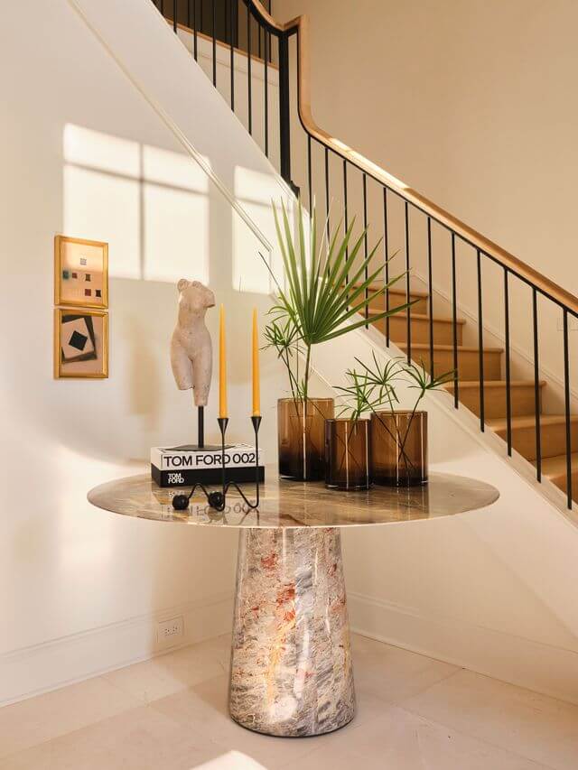 Claire Thompson's Interior Designs Are Soft, Warm, and Endlessly Livable - 14 Round marble table with decorative items, vases with palm leaves, candles, books, and a sculpture, placed in front of a staircase with black railings—Claire Thompson’s touch shines in this sunlit entryway’s warm interiors.