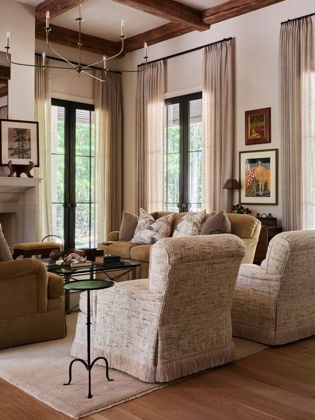 Claire Thompson's Interior Designs Are Soft, Warm, and Endlessly Livable - 3 A living room with beige upholstered furniture, a fireplace, tall windows with curtains, artwork on the wall, and wooden ceiling beams showcases warm interiors by Claire Thompson.
