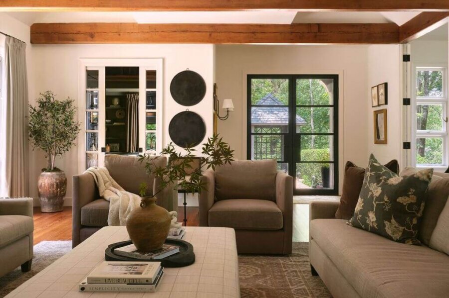 Claire Thompson's Interior Designs Are Soft, Warm, and Endlessly Livable - 7 A cozy living room with beige sofas, a large ottoman, potted plants, and wooden ceiling beams offers warm interiors, while large windows flood the textured interiors with natural light.