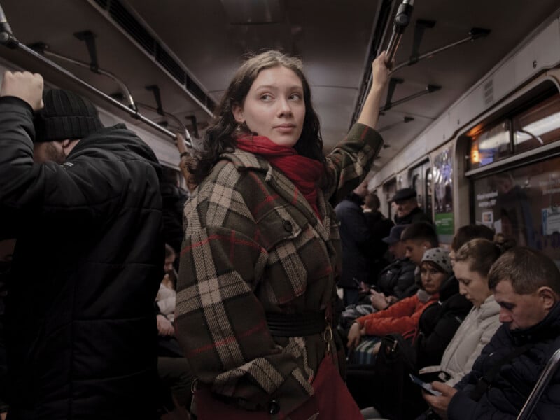 A woman in a plaid coat and red scarf stands holding a subway rail, looking to the side, surrounded by seated and standing passengers, some using their phones, in a crowded subway car.