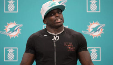 Tyreek Hill speaks at a press conference weating a white Dolphins hat and a fitted black t-shirt and a large silver chain.