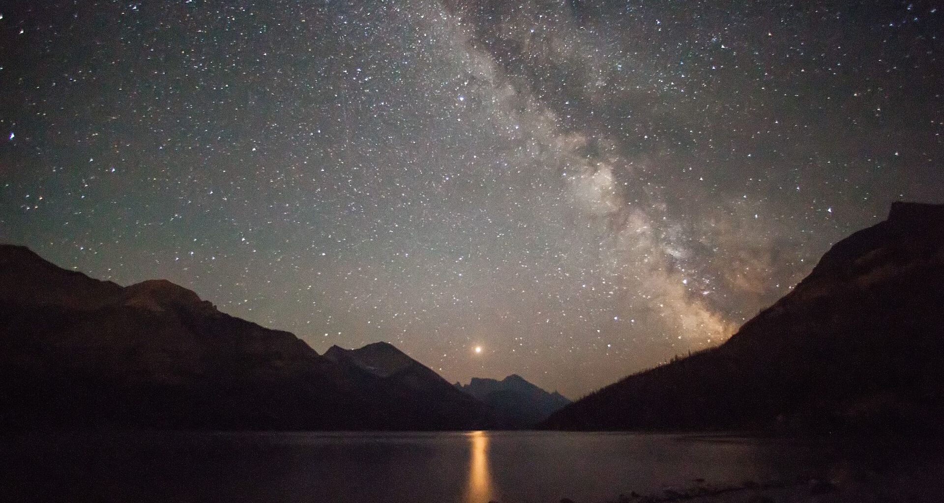 A glowing strip of the Milky Way galaxy is seen in a dark gray and purple starry night sky overlooking a lake surrounded by silhouetted jagged peaks on all sides with a glowing bright dot in the center of the image casting a yellow column of light on the dark water