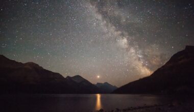 A glowing strip of the Milky Way galaxy is seen in a dark gray and purple starry night sky overlooking a lake surrounded by silhouetted jagged peaks on all sides with a glowing bright dot in the center of the image casting a yellow column of light on the dark water
