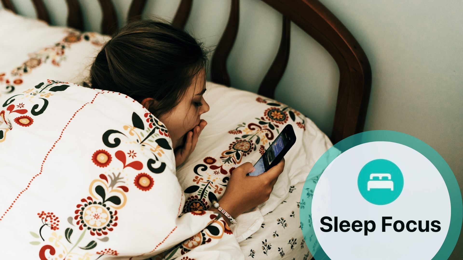 A young girl in bed with tapestry pattern sheets, laying on her front using a mobile phone as she yawns. With inset of Apple Sleep Focus logo
