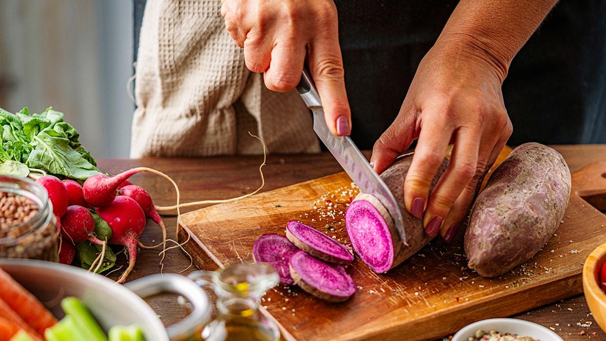 Woman slicing purple potatoes on cutting board with other produce like tomatoes and radishes around her on counter.