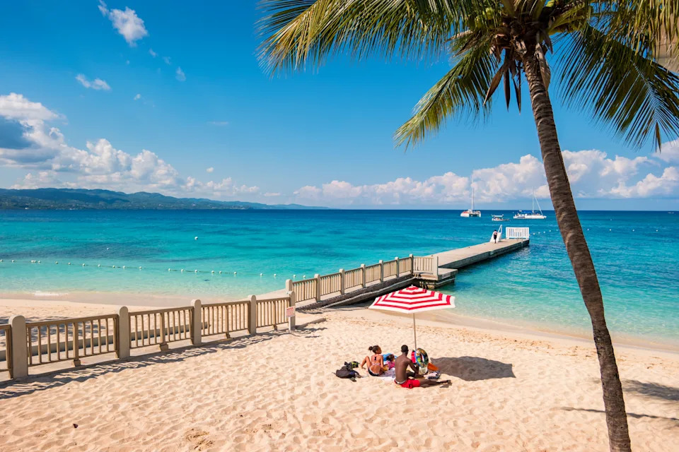 People relaxing under a beach umbrella on a sandy beach with a dock extending into the ocean and palm trees nearby