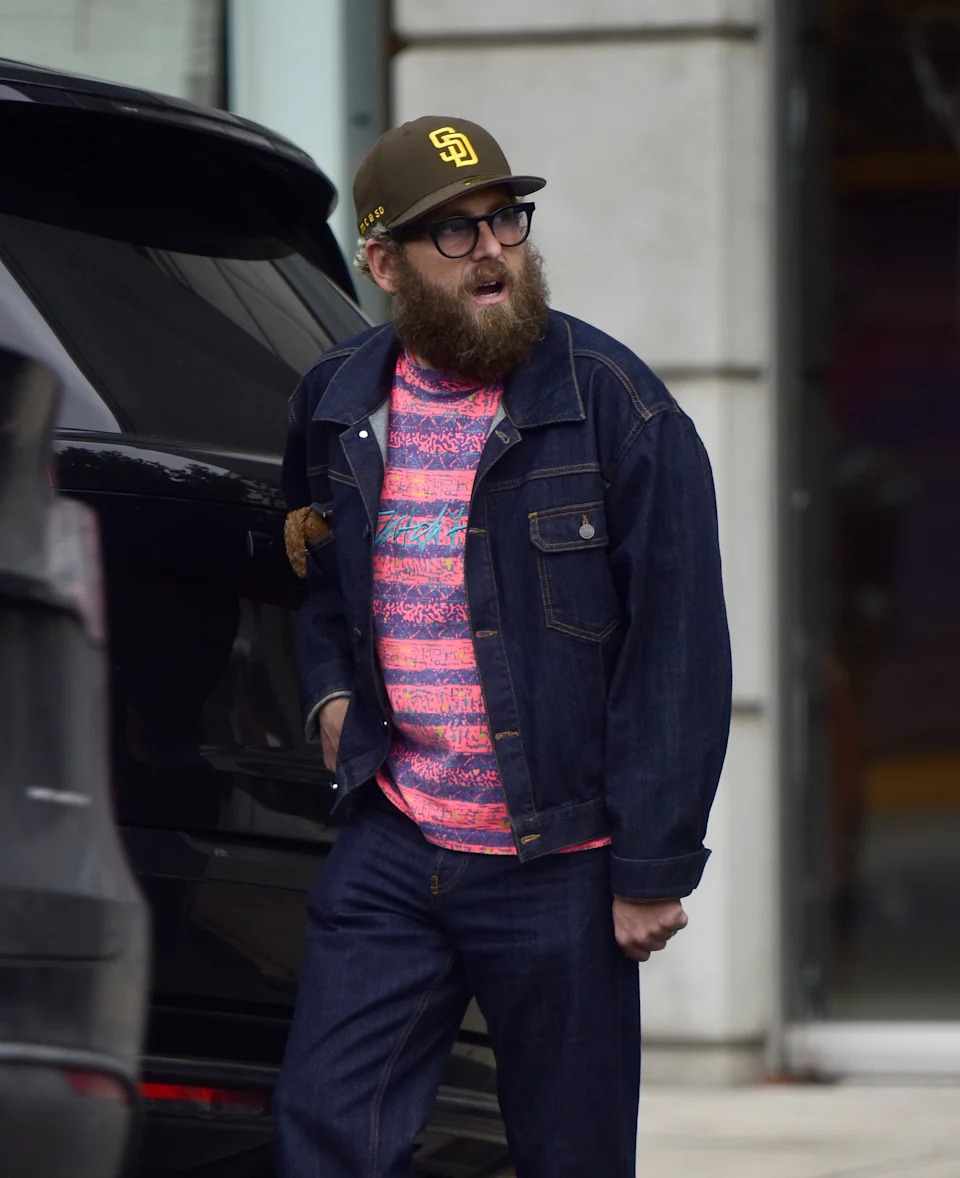 A bearded individual in glasses wears a patterned shirt, denim jacket, and pants, with a baseball cap, standing by a vehicle on a city street