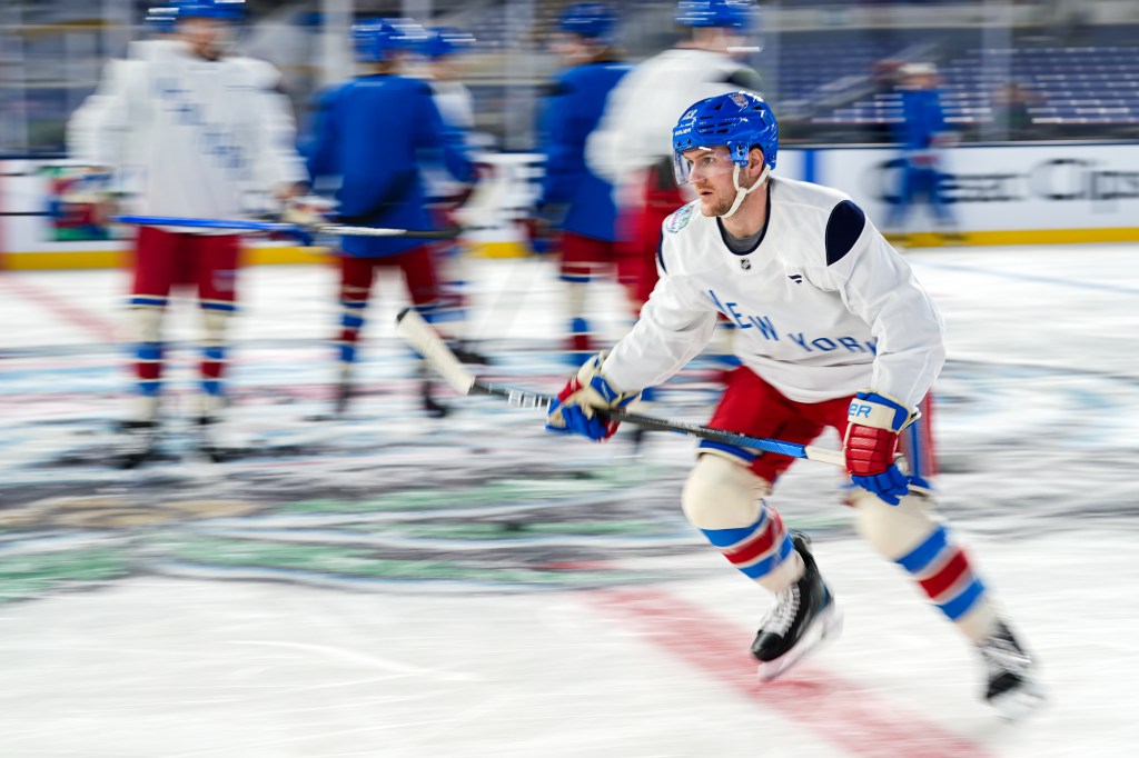 New York Rangers player Adam Fox skating across the ice with other blurred players in the background.