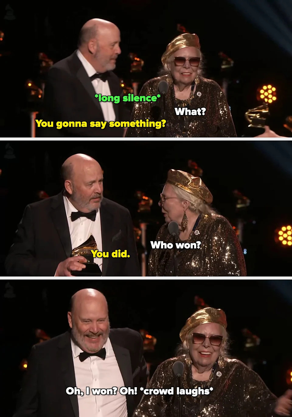 Person in glittering outfit and sunglasses, speaking on stage with a microphone, beside another person in a tuxedo