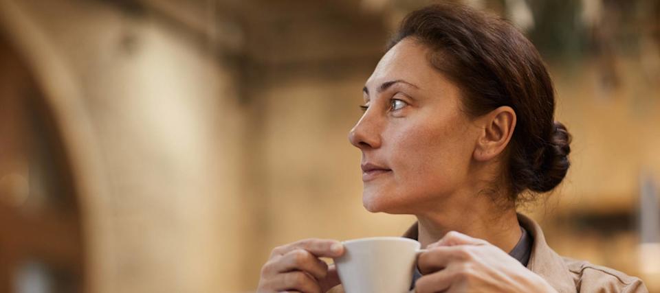 A woman sits in front of her laptop, sipping at a cup of coffee and looking into the distance.