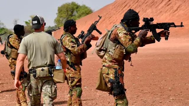 One American military trainer waka past Malian soldiers wey aim dia guns. Dem wear camouflage and dem stand in front of one hill of red earth. Dem snap di photo for 2018.