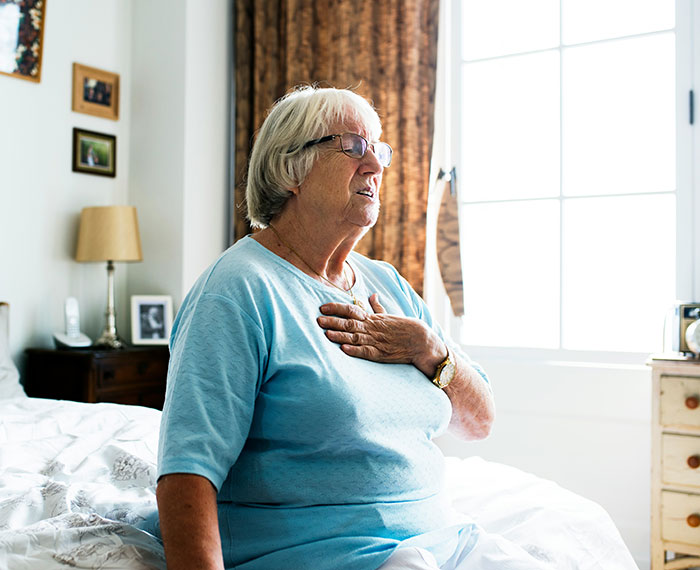 Elderly woman in glasses sitting on bed, holding chest with a funny and weirdly hilarious expression indoors.