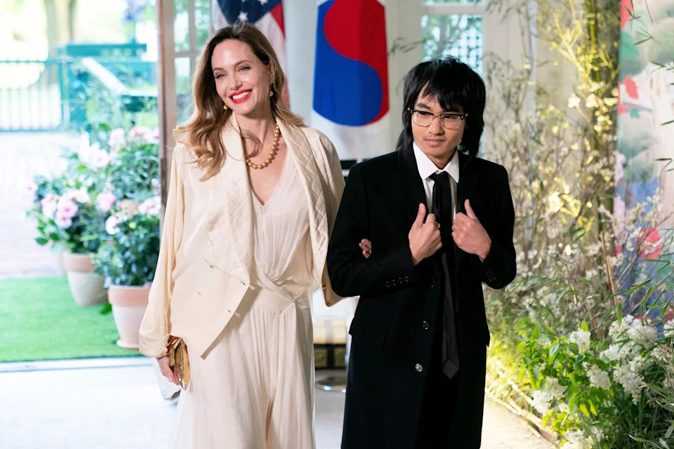 A woman in a chic cream outfit is smiling, standing arm-in-arm with a young person in a classic black suit at an elegant event