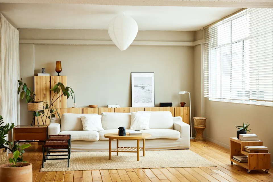 Spacious living room with a modern sofa, plants, a round wooden coffee table, and large windows letting in natural light