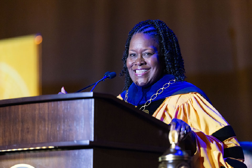 Cal State LA President stands at a podium in academic regalia, smiling while speaking during a ceremony.