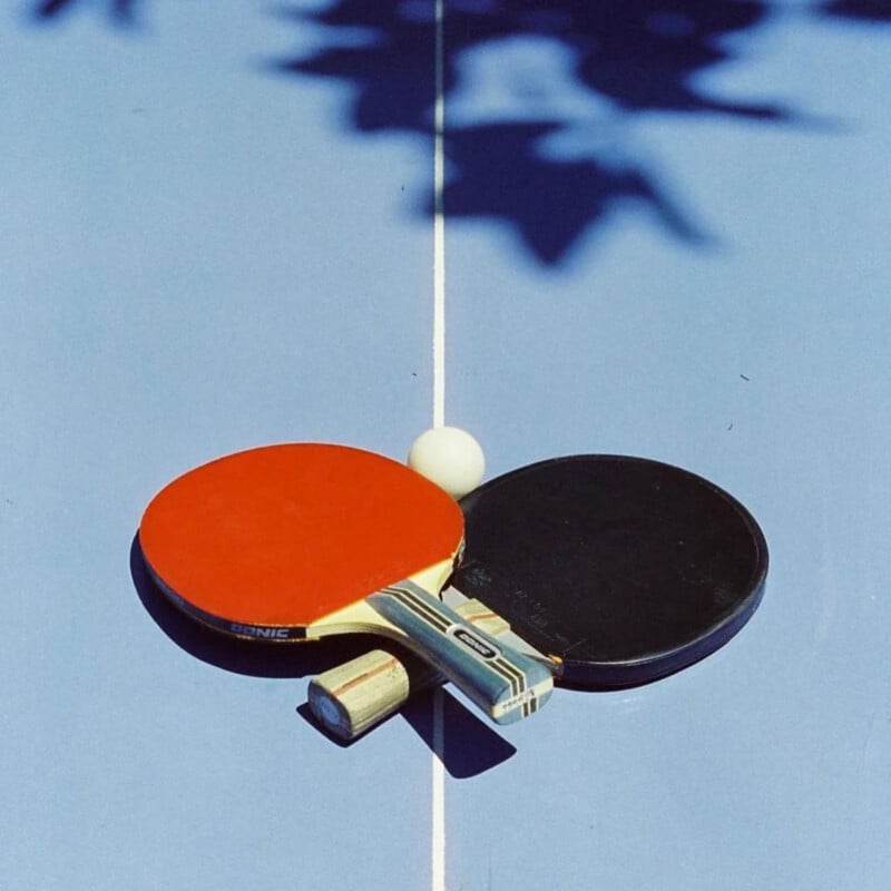 Two table tennis paddles, one red and one black, and a white ping pong ball rest on a blue ping pong table, centered over the white line. Shadow from leaves is visible on the table.